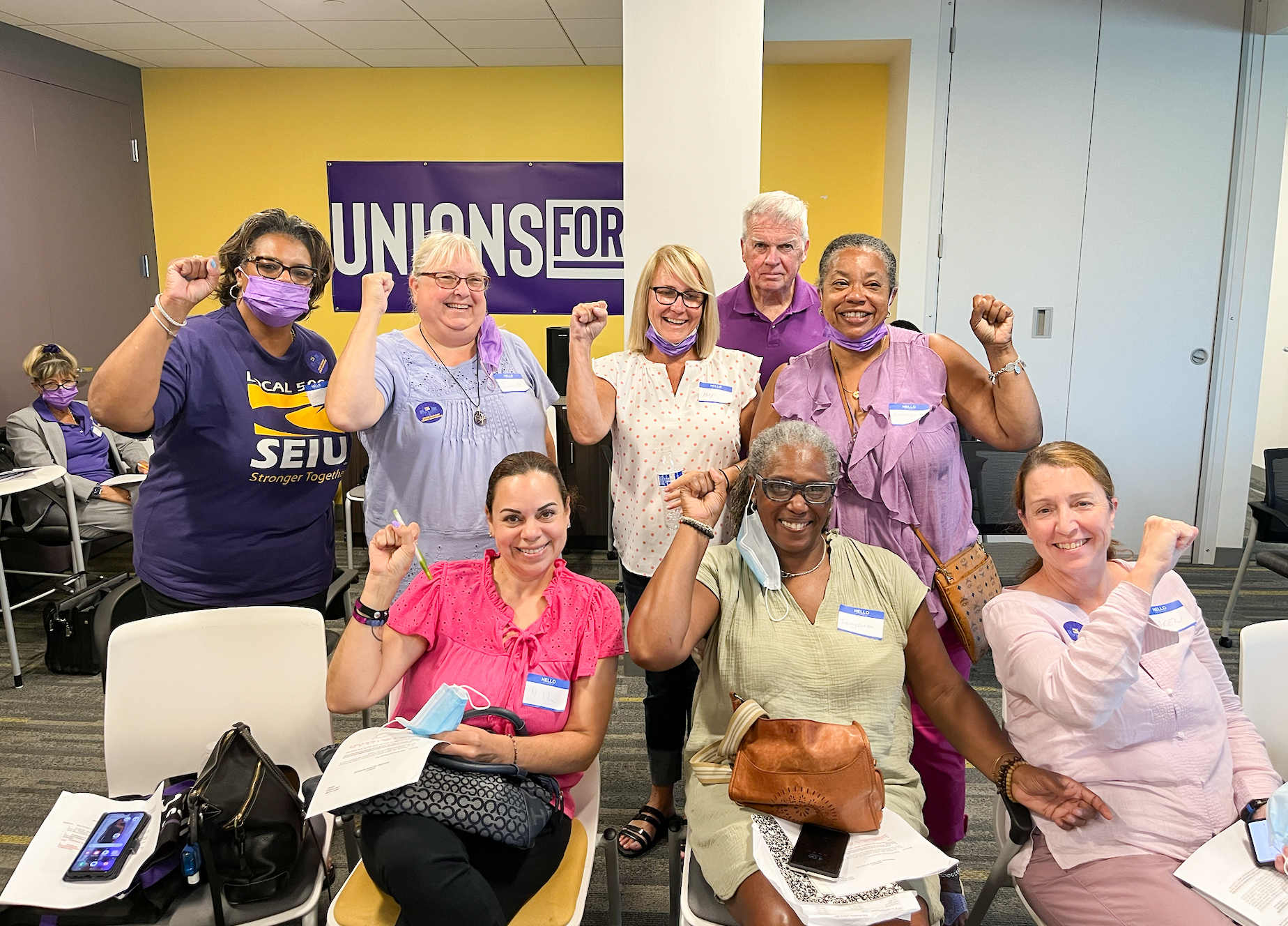 A diverse group of people, some wearing purple and union stickers, sit and stand together indoors, smiling and raising their fists in solidarity in front of a "UNIONS FOR ALL" sign.