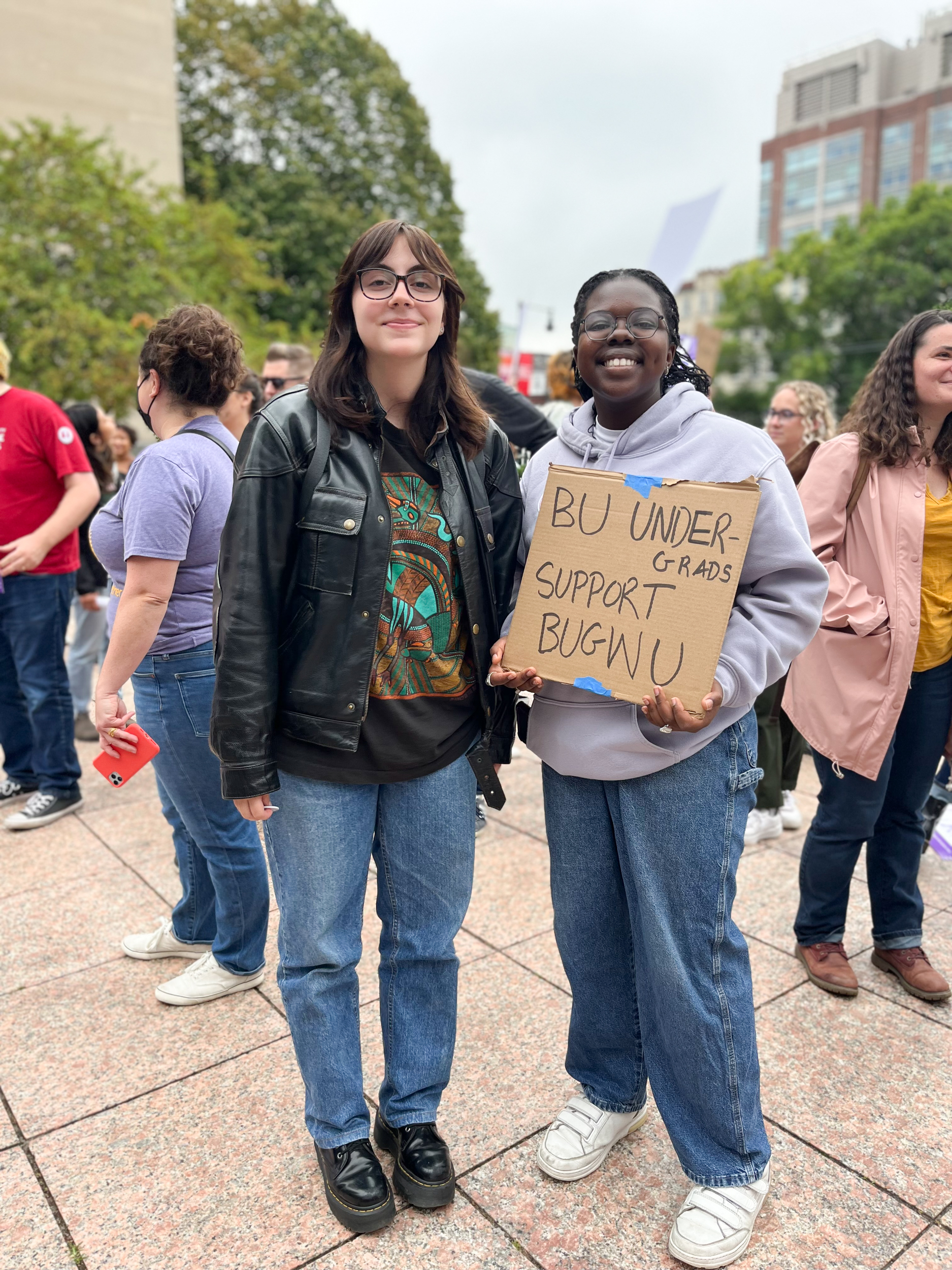 Two smiling people stand outside in a crowd. One holds a cardboard sign that says, "BU undergrads support BUGWU."