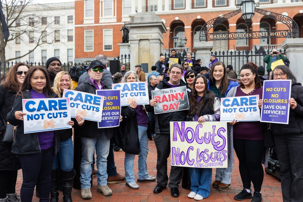 A diverse group of people stand outside the Massachusetts State House holding signs that read “Care Not Cuts,” “No Cuts No Closures,” and “No Cuts No Closures Save Our Services.”.