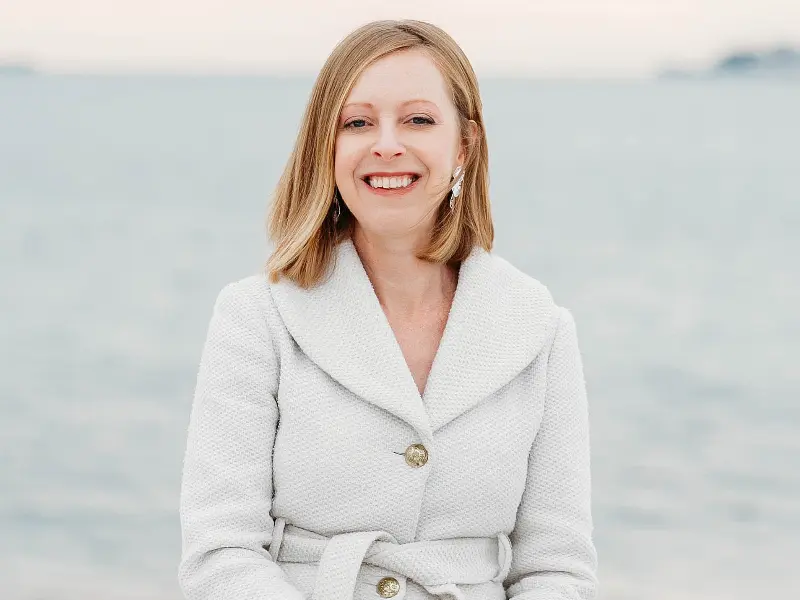 A woman with blonde hair wearing a light-colored coat smiles while sitting outdoors with a calm body of water in the background.