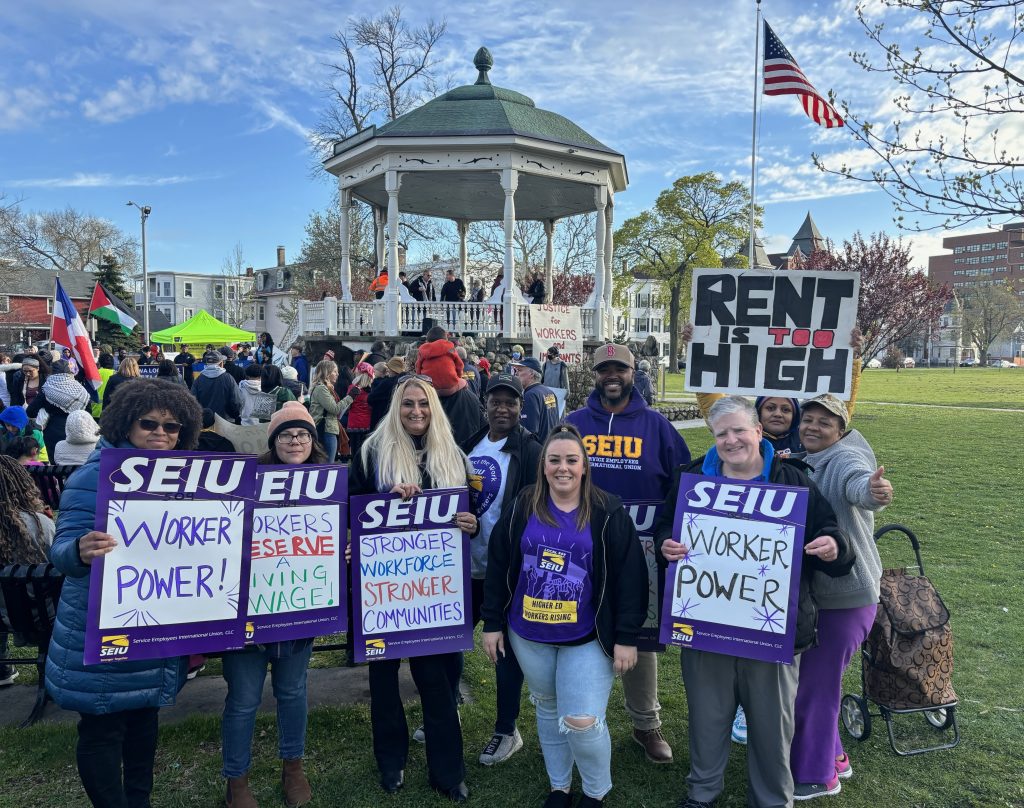 A group of people holding SEIU signs supporting worker power stands in front of a gazebo at a rally. Other people and signs, including one reading “RENT IS TOO HIGH,” are visible in the background. An American flag waves nearby.