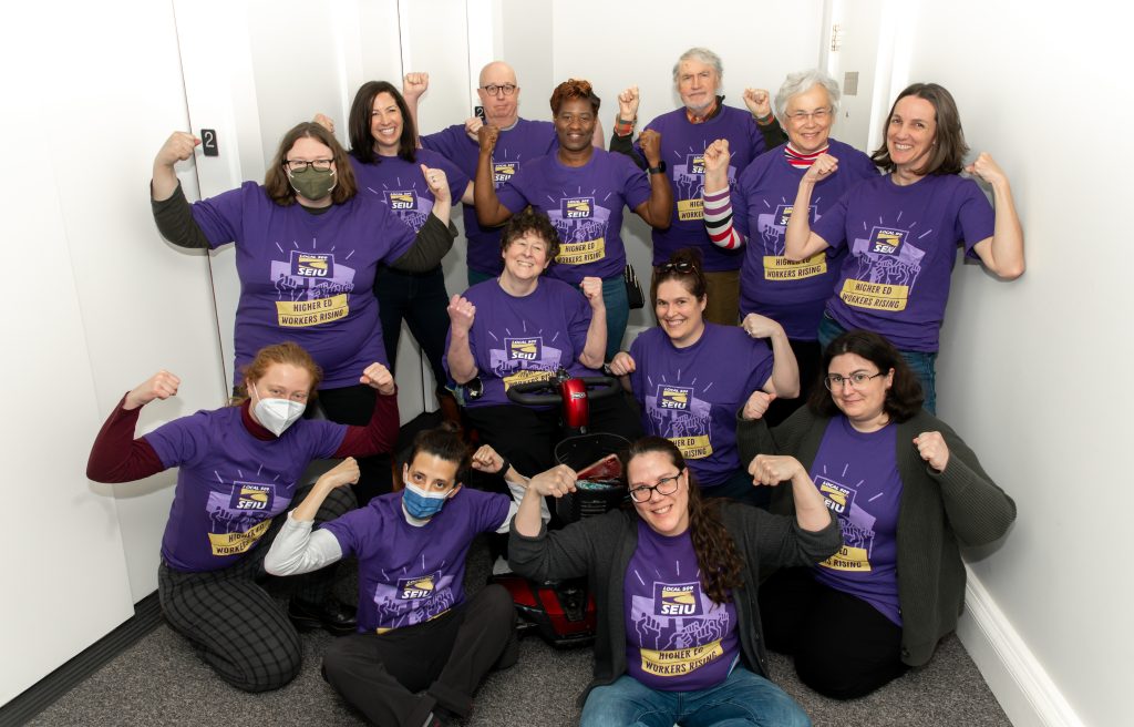A group of thirteen people wearing matching purple SEIU t-shirts pose together indoors, smiling and flexing their arms in a triumphant gesture, showing solidarity.