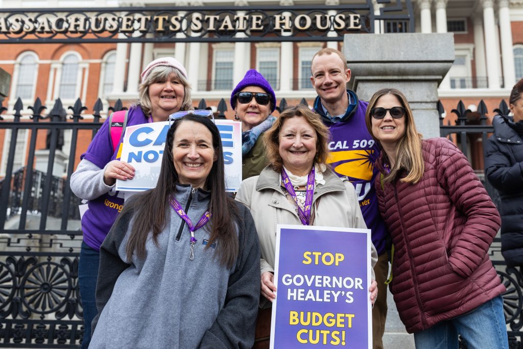 A group of smiling people stand in front of the Massachusetts State House holding protest signs, including one that says "STOP GOVERNOR HEALEY'S BUDGET CUTS!" and another reading "CARE NOT CUTS.