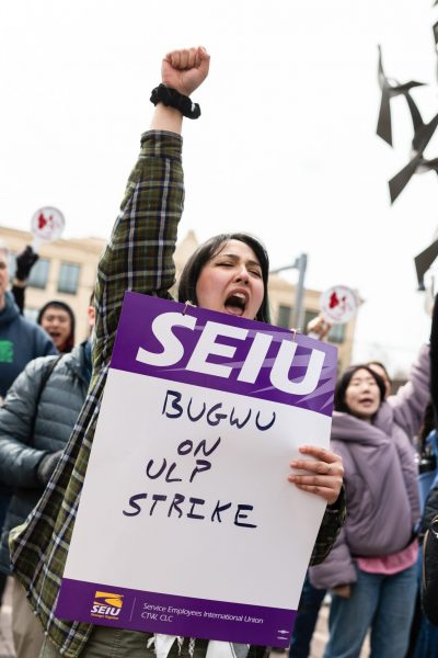 A woman holds a "SEIU BUGWU ON ULP STRIKE" sign and raises her fist while standing with a group of demonstrators at an outdoor protest.
