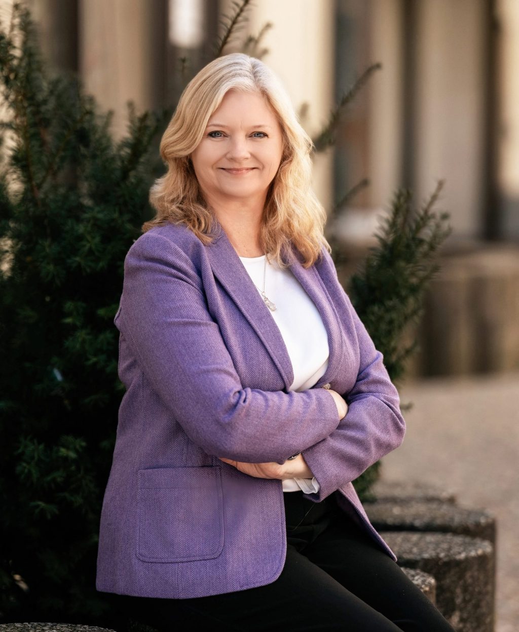A woman with blonde hair wearing a purple blazer and white top sits outdoors, smiling with arms crossed. There are green bushes and a building in the background.
